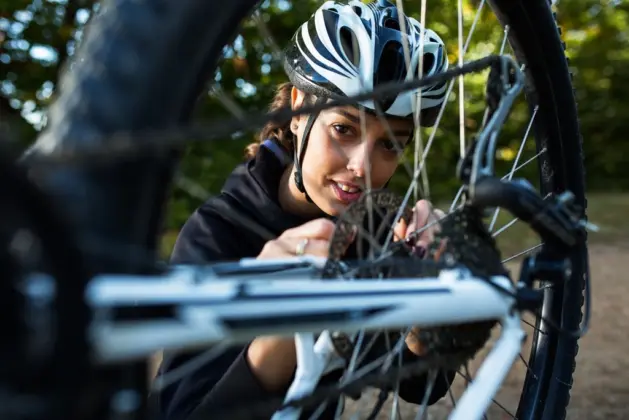 Estudiante universitario realizando mantenimiento de bicicleta en un parque, revisando la cadena con un trapo y lubricante, bicicleta urbana de color azul, fondo con edificios de campus