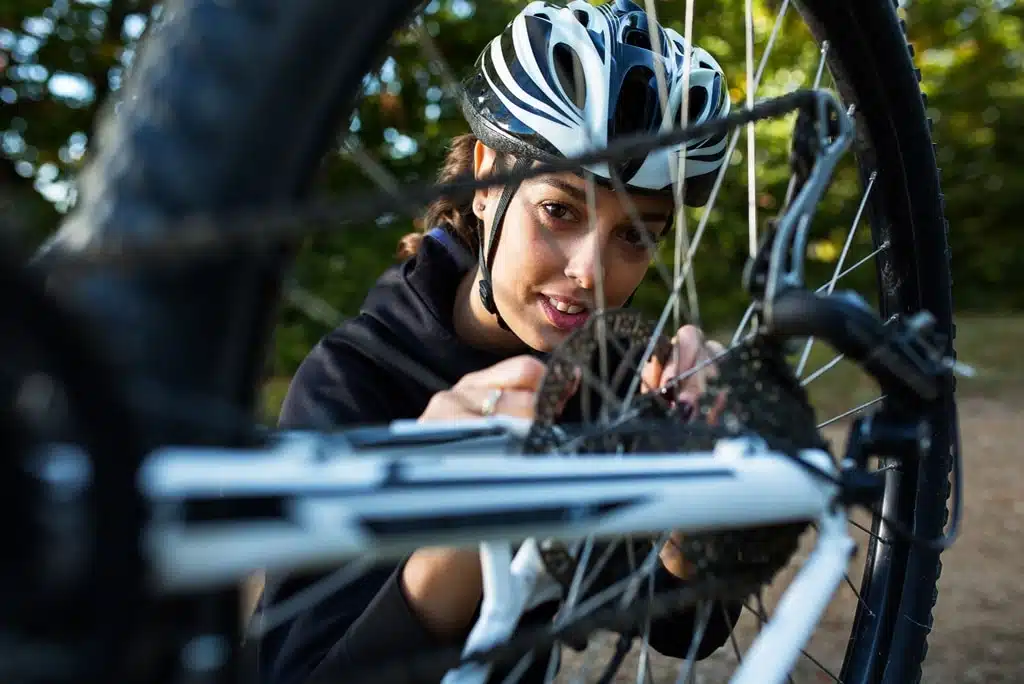 Estudiante universitario realizando mantenimiento de bicicleta en un parque, revisando la cadena con un trapo y lubricante, bicicleta urbana de color azul, fondo con edificios de campus