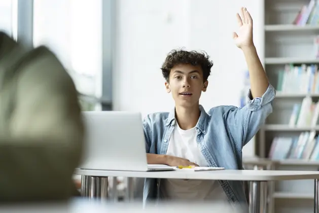 Joven en salón de clase levantando la mano para hacer una pregunta inteligente en una reunión de equipo, demostrando confianza y comunicación efectiva.