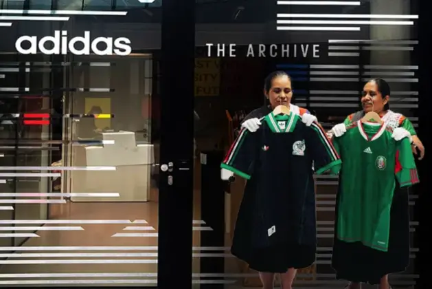 Artesanas mexicanas con guantes blancos colocando el jersey de México en El Archivo de Adidas sobre una vitrina iluminada, fondo de estanterías metálicas con camisetas históricas.