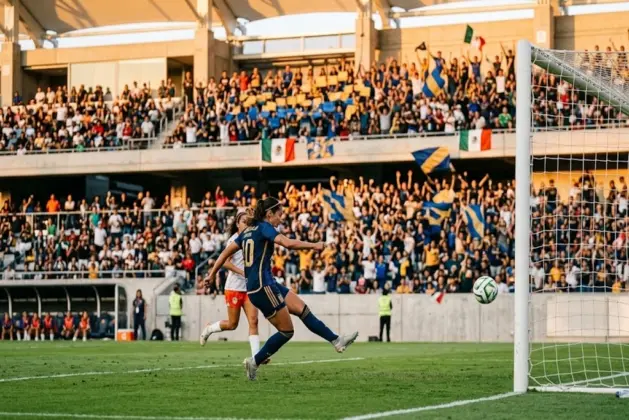Jugadoras de fútbol femenil en acción durante un partido histórico en un estadio lleno de aficionados jóvenes, con camisetas de equipos mexicanos, celebrando un gol bajo luz natural.
