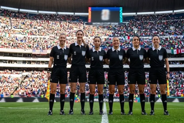 Seis mujeres árbitras Mundial posan con sus silbatos en el centro del campo, luciendo los uniformes oficiales del torneo, con el estadio lleno de aficionados al fondo y la bandera del Mundial ondeando.