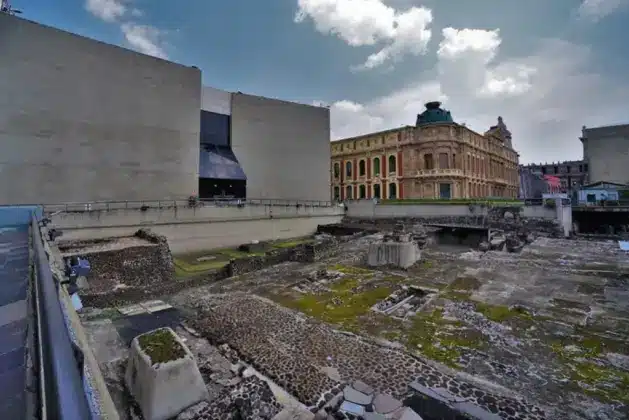 Vista frontal del basamento del Templo de Ehécatl y los muros del Juego de Pelota en el Museo del Templo Mayor de la CDMX, con detalles de la piedra volcánica y el contexto urbano del centro histórico.