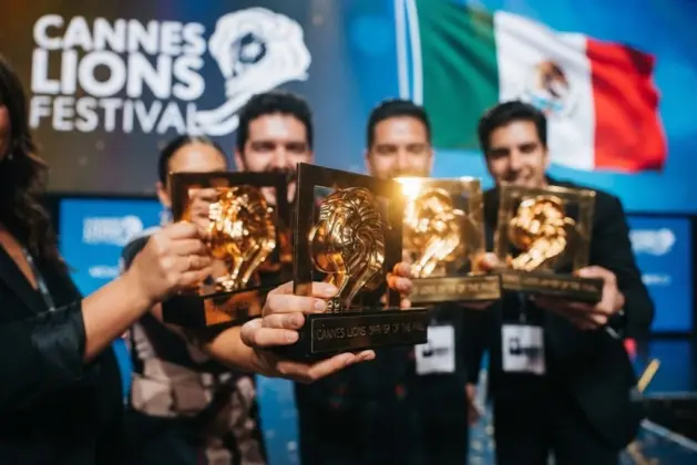 Grupo de creativos mexicanos celebrando en el escenario del festival Cannes Lions con estatuillas doradas en mano, fondo con la pantalla del evento y la palabra "México" proyectada